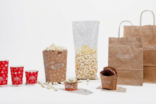 En samling take away-förpackningar från Gröna Lund uppställda mot en vit bakgrund. Bilden visar rödvita mönstrade muggar, popcornbägare i papp, en genomskinlig popcornpåse, en donutask med en chokladdonut, samt bruna papperskassar med vit Gröna Lund-logotyp.