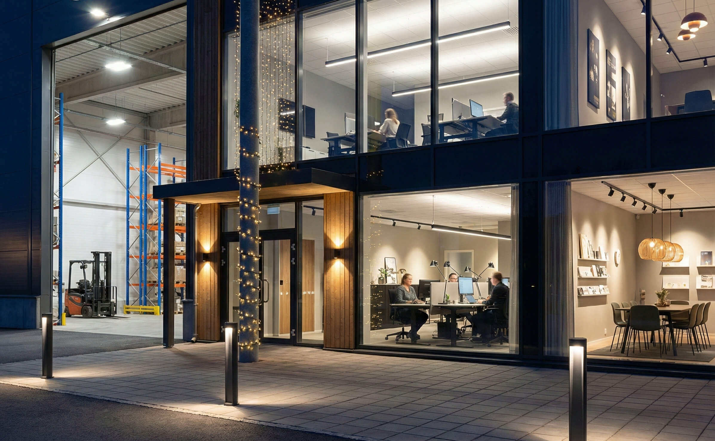 LjuslingorKvällsbild av en modern fastighet som visar olika belysningsprodukter installerade. I förgrunden syns tända pollare längs en gångväg och väggarmaturer som lyser upp fasaden. Inne i kontorsdelen till höger syns nedpendlade linjära armaturer, infällda takpaneler samt en del dekorativ belysning. Till vänster genom en stor port syns ett lager upplyst av kraftiga, höghängande industriarmaturer (high-bay).