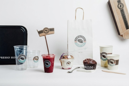 Ett sortiment av mat och dryck för take away från Espresso House mot en vit bakgrund. I mitten står en röd smoothie med sugrör och flagga, omgiven av en yoghurtbägare med granola, en chokladmuffin, kaffemuggar i olika storlekar, en vit papperskasse samt en svart bricka.