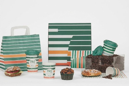 En uppsättning 7-Eleven-produkter med deras karakteristiska randiga mönster i grönt, vitt och orange. Bilden inkluderar en papperskasse, kaffemuggar med texten "Hej", en smörgås, en muffins, en lussekatt och en genomskinlig låda fylld med kaffebönor.