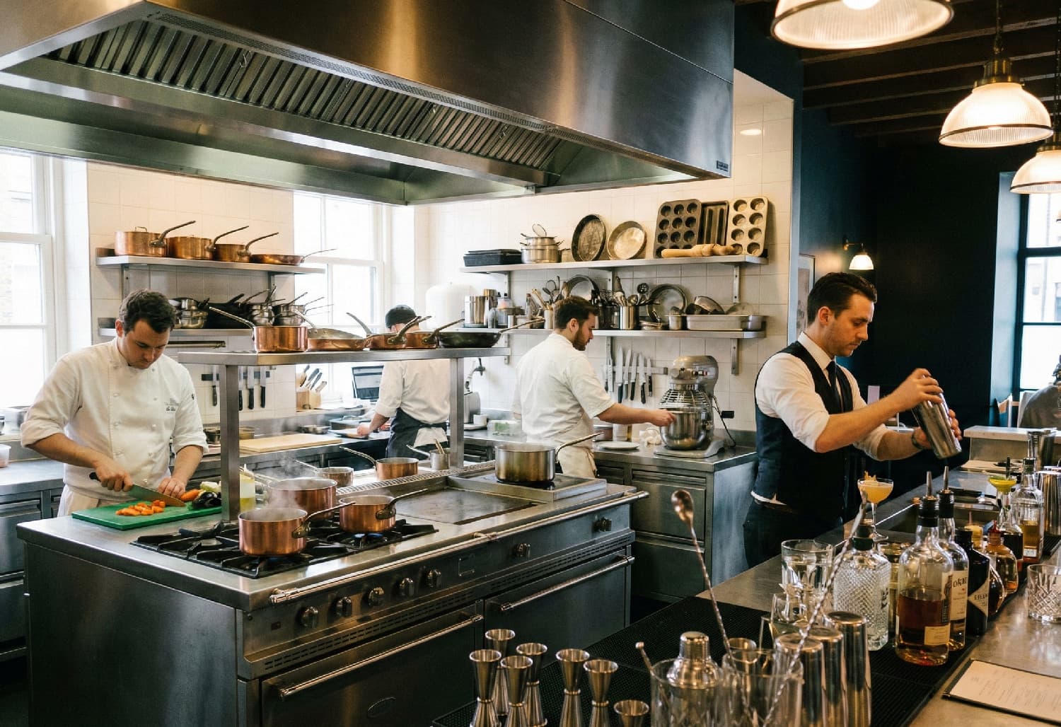 Professionellt restaurangkök och bar i full aktivitet. Kockar förbereder mat vid en rostfri köksö med kopparkastruller medan en bartender skakar en drink omgiven av barutrustning.