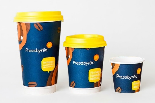 Tre kaffemuggar i papp från Pressbyrån i storlekarna stor, mellan och liten. Muggarna är mörkblå med gula lock och orangea grafiska mönster samt vit text som lyder "Pressbyrån"