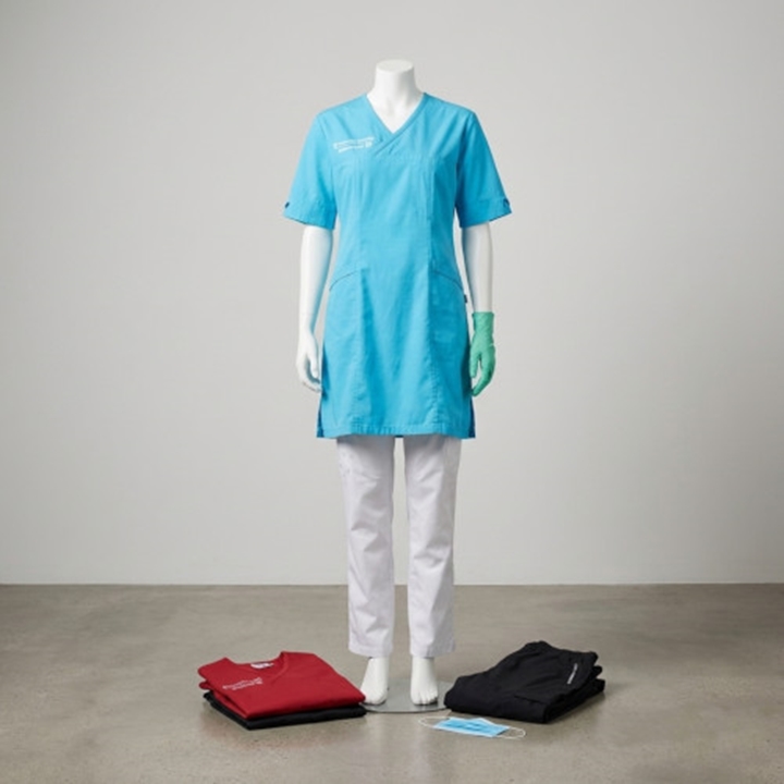 Funktionella och bekväma vårdkläder med tryckmöjligheter. Bilden visar en ljusblå bussarong med klinikens logotyp och vita vårdbyxor på en provdocka. Intill ligger vikta alternativ i rött och svart, vilket visar bredden av färger för hemtjänst, tandvård och sjukhuspersonal.