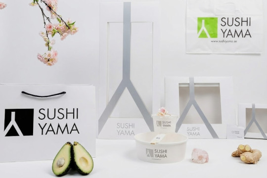 Stilrena vita take away-förpackningar från Sushi Yama med minimalistisk svart och silverfärgad logotyp. Bilden visar papperskassar, rektangulära sushilådor med fönster och en rund skål. Färsk avokado, ingefära och en kvist körsbärsblommor ligger som dekoration bredvid.