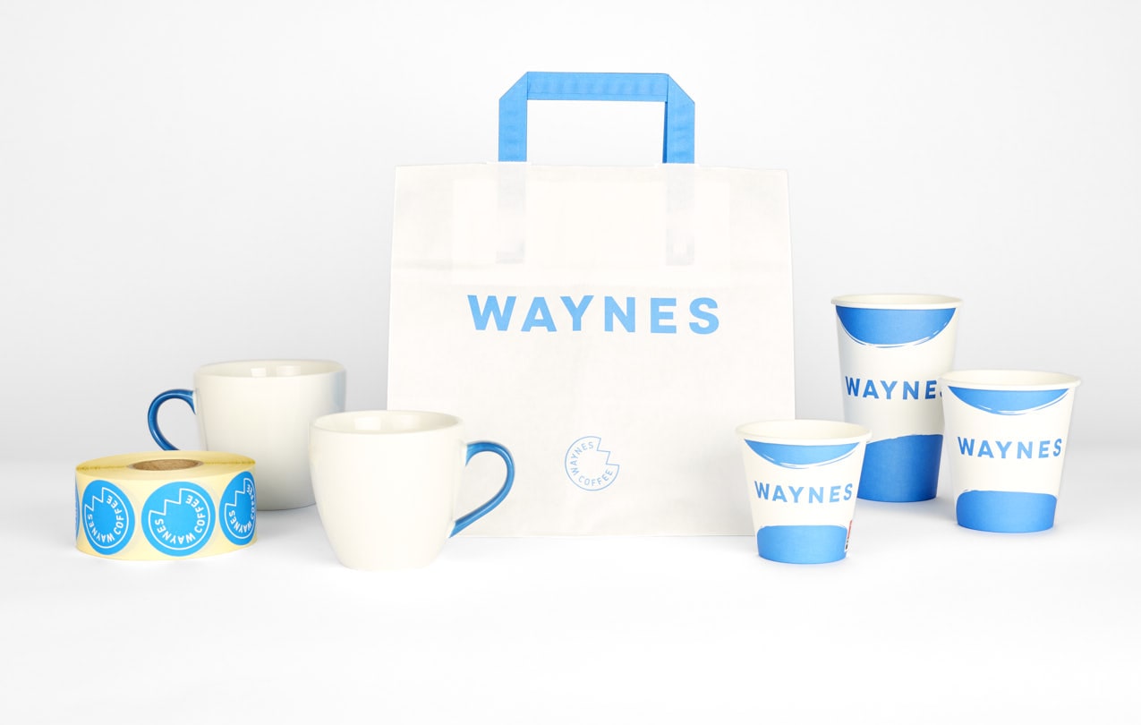 Varumärkesprofilering för Waynes Coffee i vitt och blått. Bilden innehåller en vit papperskasse med blå logotyp och handtag, keramiska muggar med blå detaljer, take away-muggar i papp med blått penseldragsmönster samt en rulle med runda klistermärken.