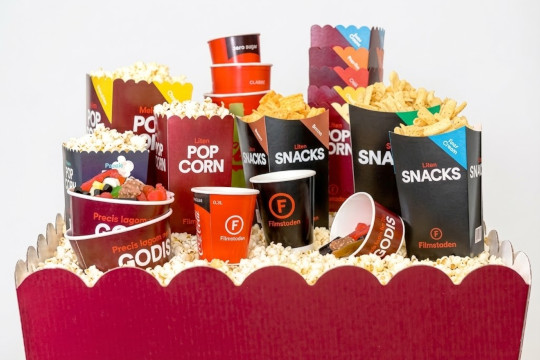En stor röd popcorn-formad behållare fylld med popcorn, som i sin tur rymmer en mängd olika Filmstaden-förpackningar. Där finns popcornbägare, svarta snackslådor för nachos, röda bägare för godis samt staplade dryckesmuggar i rött och svart.